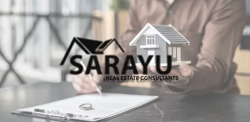 Person holds a small house model over a desk with a clipboard featuring the Sarayu Real Estate Consultants logo